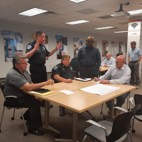 A group of public safety officials and staff meet around conference tables in a training room. One uniformed police officer stands and gestures while speaking to the group, as others—including another uniformed officer and several seated participants taking notes—listen and review documents spread across the table.
