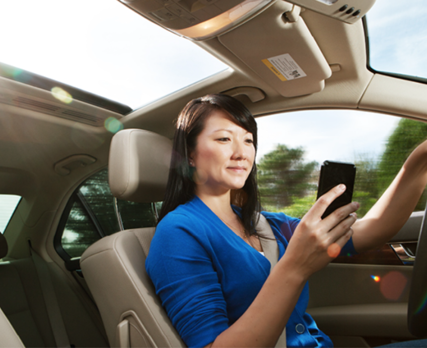 A woman sitting in the driver's seat of a moving car looking down at a smartphone in her hands.