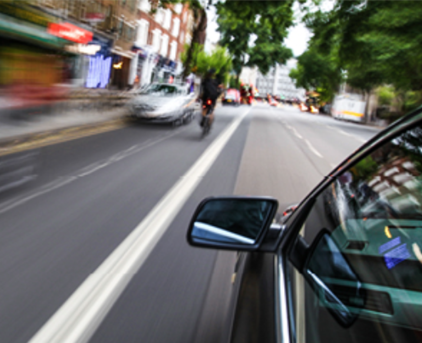 View from inside a speeding car driving down a tree-lined city street, showing the road ahead and the side mirror.