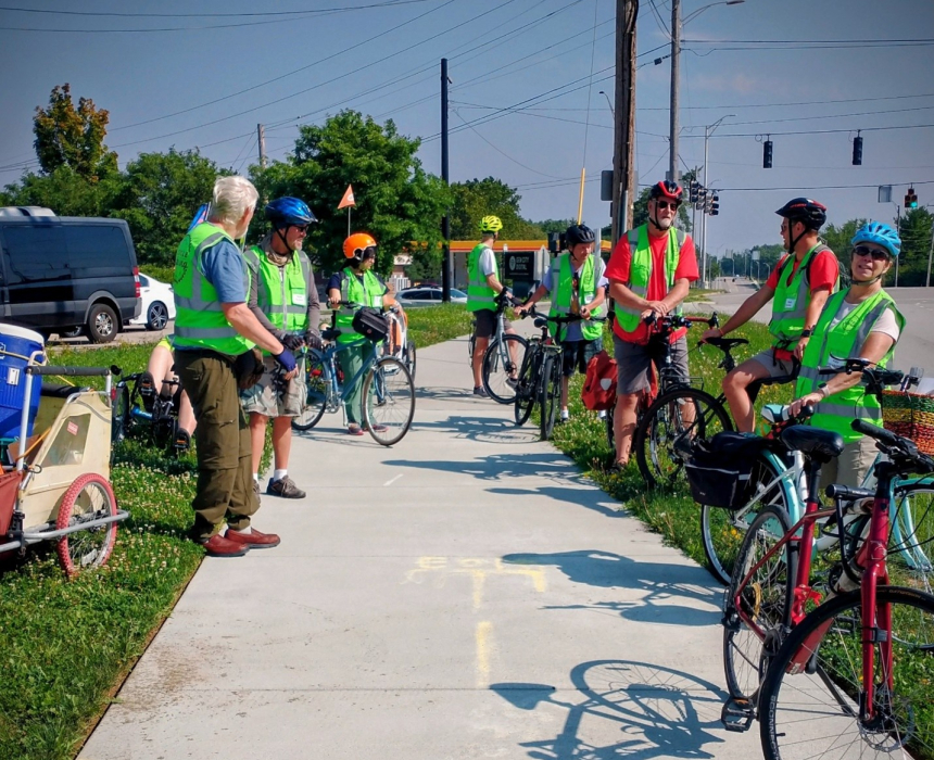 A group of cyclists and pedestrians wearing high-visibility safety vests and helmets gathered on a paved sidewalk next to a roadway.
