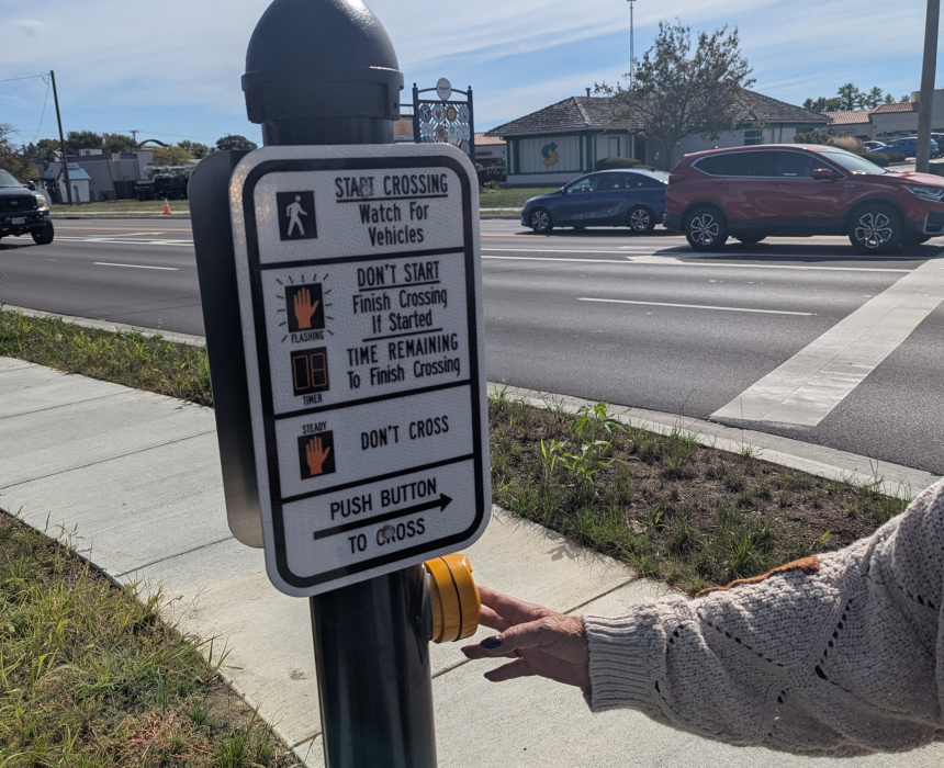 Person pressing a pedestrian crossing button beside a multi-lane road, with traffic passing through the intersection.