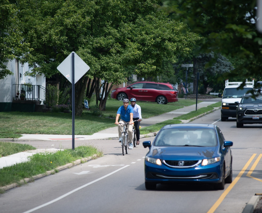 Two cyclists riding in a marked bike lane alongside vehicle traffic on a tree-lined residential street.