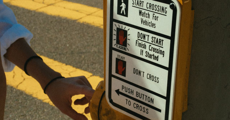 Person pressing crosswalk button on sunny day for street crossing.
