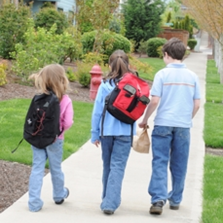 Three children wearing backpacks walk together along a neighborhood sidewalk on their way to school.