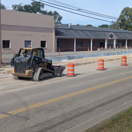 Road construction along Main Street in Fairborn, featuring a John Deere track loader, orange traffic barrels, and new concrete curbing in front of the local Eagles building.