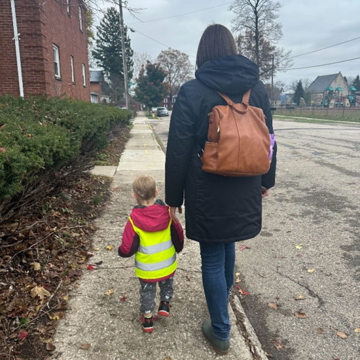 An adult and a small child walk on a sidewalk next to a parking lot