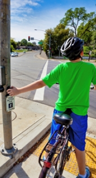A child wearing a bicycle helmet waits at a crosswalk and presses the pedestrian crossing button before crossing the street.